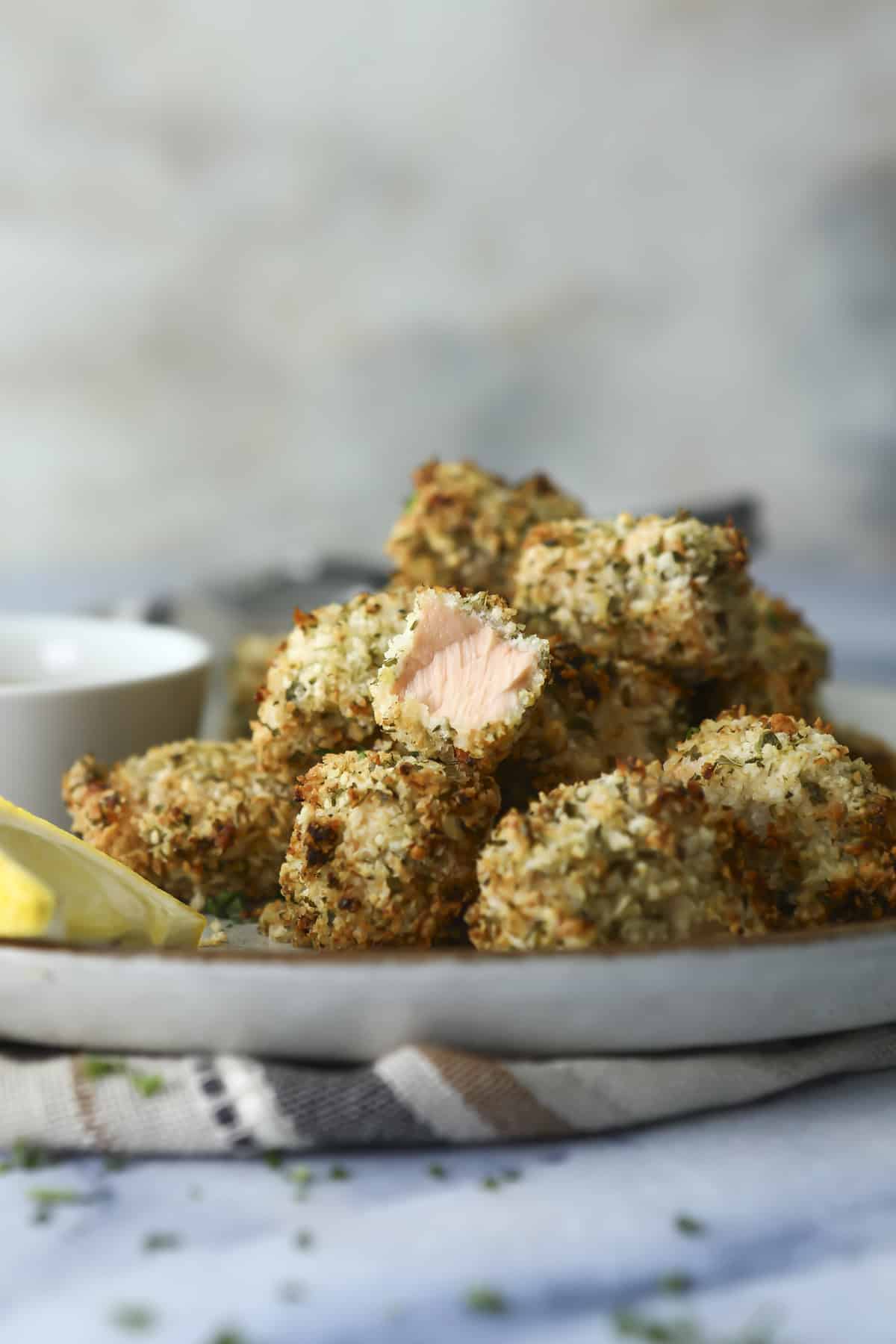 A plate of crispy, breaded salmon bites is shown. One piece is bitten to reveal the pink salmon inside. Lemon wedges and a small bowl of dipping sauce are on the side. The background is softly blurred.