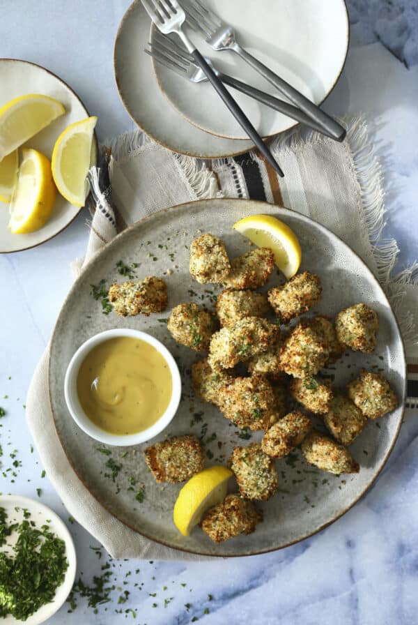 A plate of breaded, baked bite-sized pieces served with lemon wedges and a bowl of dipping sauce, placed on a cloth napkin with plates, forks, and extra lemon slices nearby. Fresh herbs are sprinkled on the food and around the plate. Baked Salmon Bites