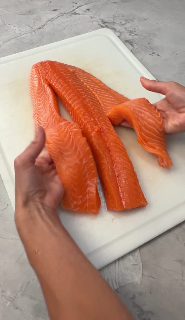 Two hands hold and arrange large, fresh, orange salmon fillets on a white cutting board, which sits on a light gray countertop.