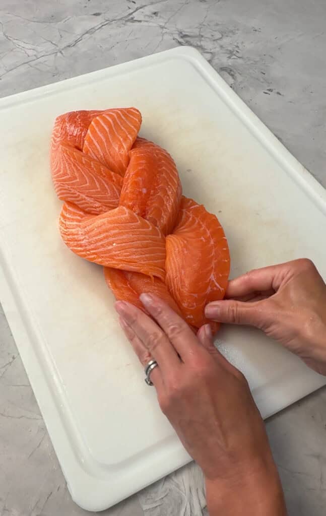 Two hands are braiding three strips of raw salmon on a white cutting board placed on a gray marble surface.