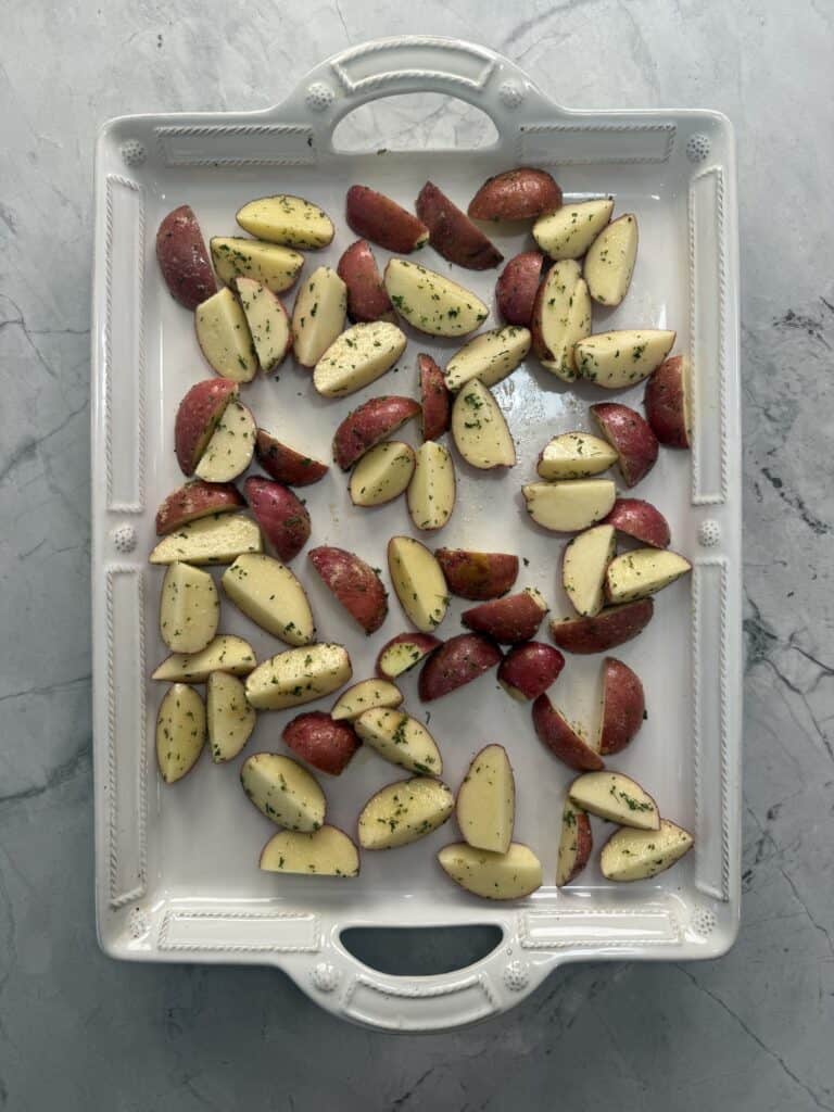 Quartered red potatoes seasoned with herbs, spread evenly on a white rectangular ceramic baking dish, set on a gray marble surface.