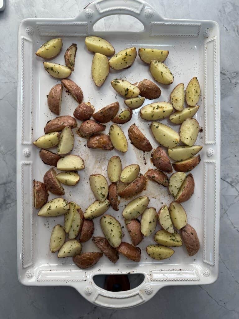 A white rectangular tray with roasted, sliced red potatoes sprinkled with herbs, spread out evenly on a marble countertop.
