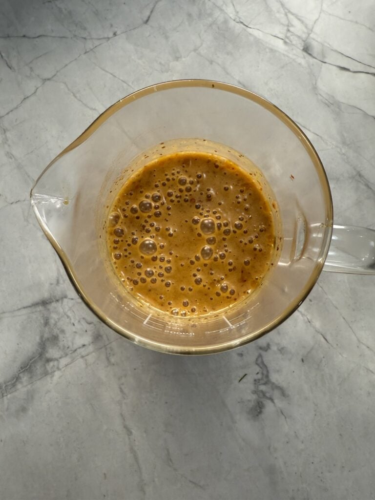 A glass measuring cup filled with a frothy orange sauce or dressing sits on a light gray marble countertop.