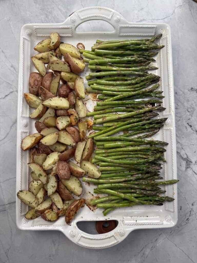 A white rectangular baking dish holds roasted red potato wedges on the left and roasted asparagus spears on the right, all seasoned with herbs, on a marble countertop.