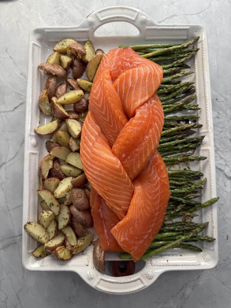 A white tray with a braid of raw salmon fillets in the center, roasted potato wedges on the left, and seasoned asparagus spears on the right, placed on a gray marble surface.
