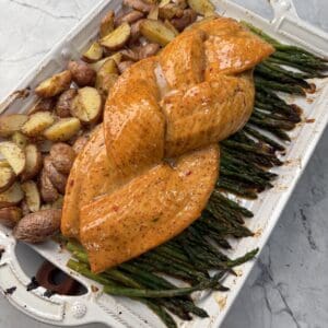A white baking dish with seasoned, braided Honey mustard salmon on a bed of asparagus, next to roasted potatoes, on a light gray marble surface.