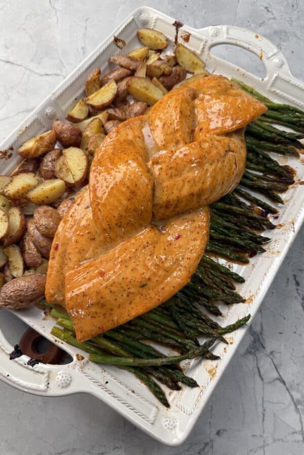 A white baking dish with seasoned, braided Honey mustard salmon on a bed of asparagus, next to roasted potatoes, on a light gray marble surface.
