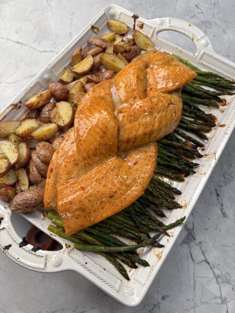 A white baking dish with seasoned, braided Honey mustard salmon on a bed of asparagus, next to roasted potatoes, on a light gray marble surface.