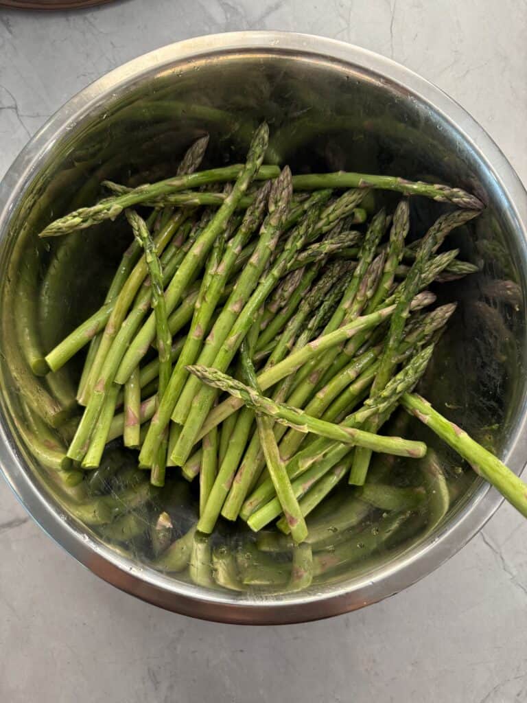 A stainless steel bowl filled with fresh, uncooked asparagus spears sits on a gray countertop.