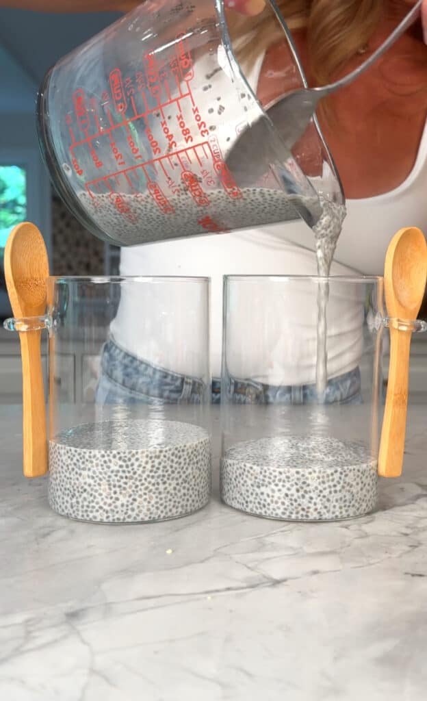 A person pours chia seed pudding from a glass measuring cup into two clear glasses on a marble counter, each with a wooden spoon attached.