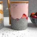 A glass jar filled with high protein chia pudding, sliced strawberries, and pink yogurt, topped with a wooden lid and spoon. Next to it is part of another jar and a bowl of fresh strawberries on a white surface.