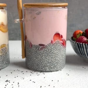 A glass jar filled with high protein chia pudding, sliced strawberries, and pink yogurt, topped with a wooden lid and spoon. Next to it is part of another jar and a bowl of fresh strawberries on a white surface.