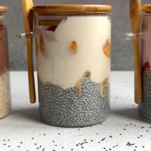 A glass jar filled with high protein banana cream chia pudding, yogurt, and fruit, topped with a wooden lid and matching spoon, sits on a white surface with chia seeds scattered nearby.