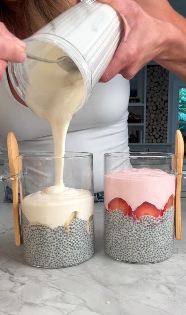 A person pours a creamy mixture into a glass containing chia pudding and banana slices; next to it is another glass with chia pudding, strawberry slices, and a layer of pink cream. Both glasses have small wooden spoons.