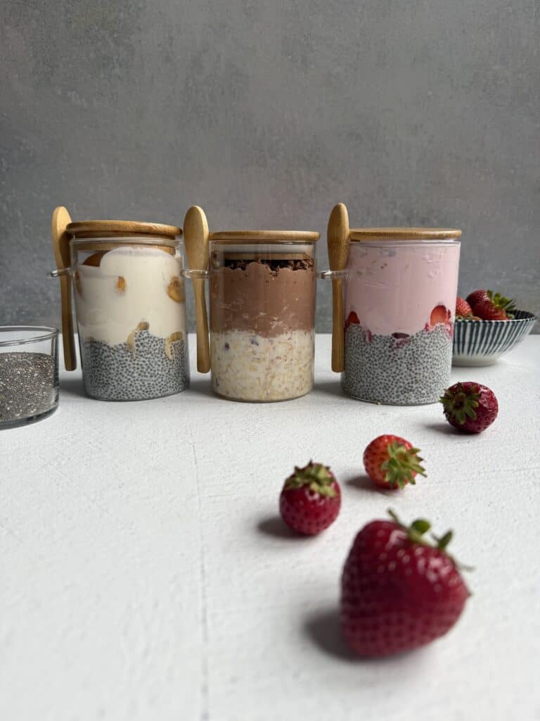 Three jars of high protein chia pudding are arranged in a row, each topped with different ingredients. Wooden spoons rest on the jars. Fresh strawberries and a bowl of strawberries are nearby on a white surface.