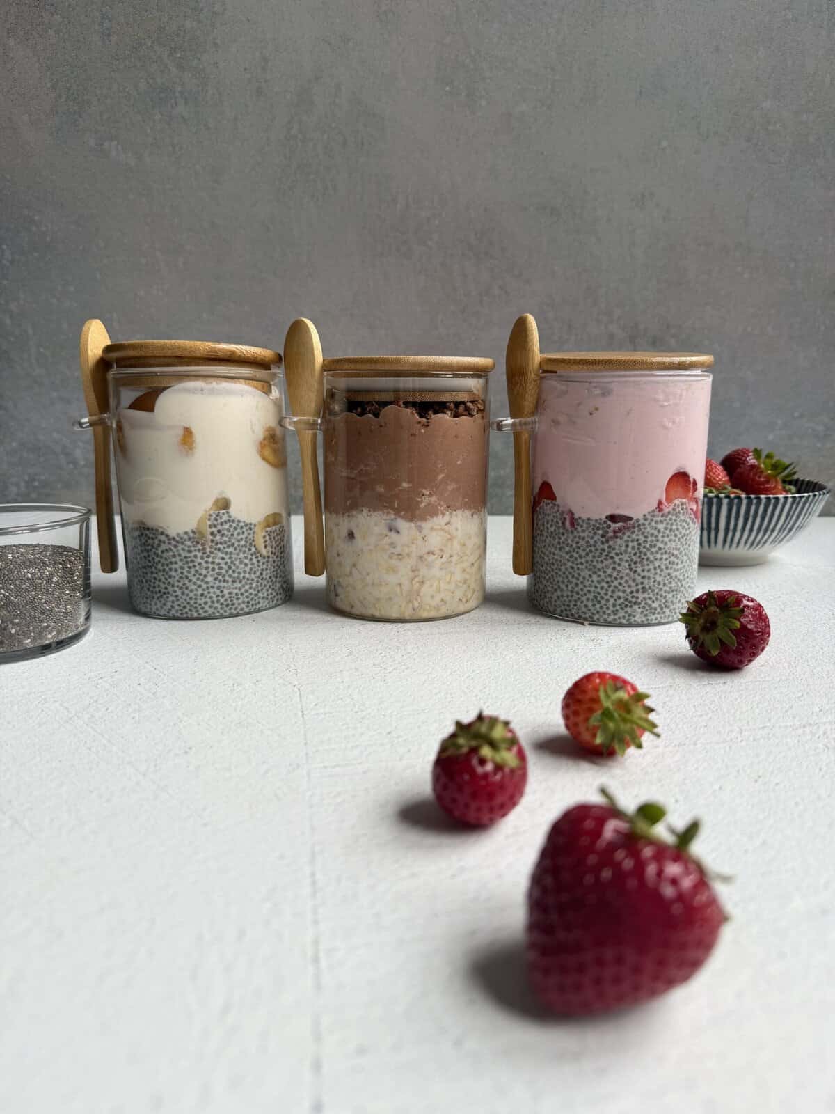 Three jars of high protein chia pudding are arranged in a row, each topped with different ingredients. Wooden spoons rest on the jars. Fresh strawberries and a bowl of strawberries are nearby on a white surface.