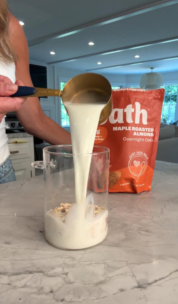 A person pours milk from a brass measuring cup into a glass jar containing oats on a kitchen counter. An orange bag labeled “Oath Maple Roasted Almond Overnight Oats” is in the background.