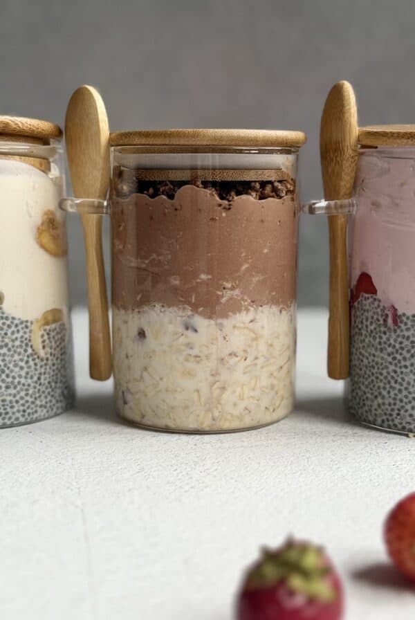 Three glass jars with bamboo lids and spoons, each containing layered high protein overnight oats with chia pudding, yogurt, and fruit. The middle jar has chocolate yogurt, and strawberries are visible in front.