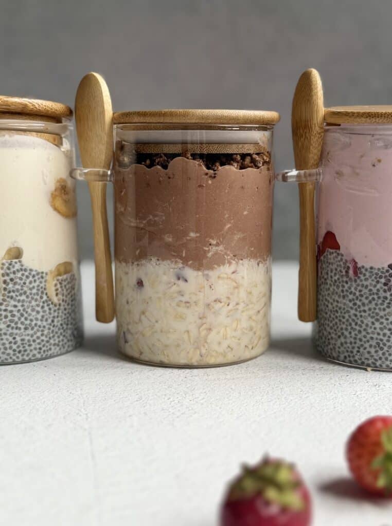 Three glass jars with bamboo lids and spoons, each containing layered high protein overnight oats with chia pudding, yogurt, and fruit. The middle jar has chocolate yogurt, and strawberries are visible in front.