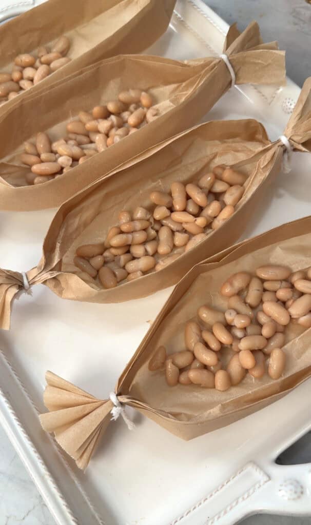 Three small boats made from brown parchment paper, each filled with cooked white beans, are tied at the ends with white string and arranged on a white tray.