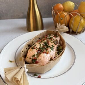 A piece of baked salmon garnished with herbs and red peppers, served in parchment paper on a white plate. In the background, a gold vase and a basket of oranges and lemons are visible.