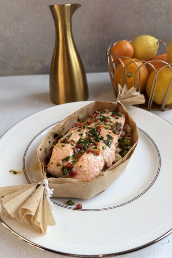 A piece of baked salmon garnished with herbs and red peppers, served in parchment paper on a white plate. In the background, a gold vase and a basket of oranges and lemons are visible.