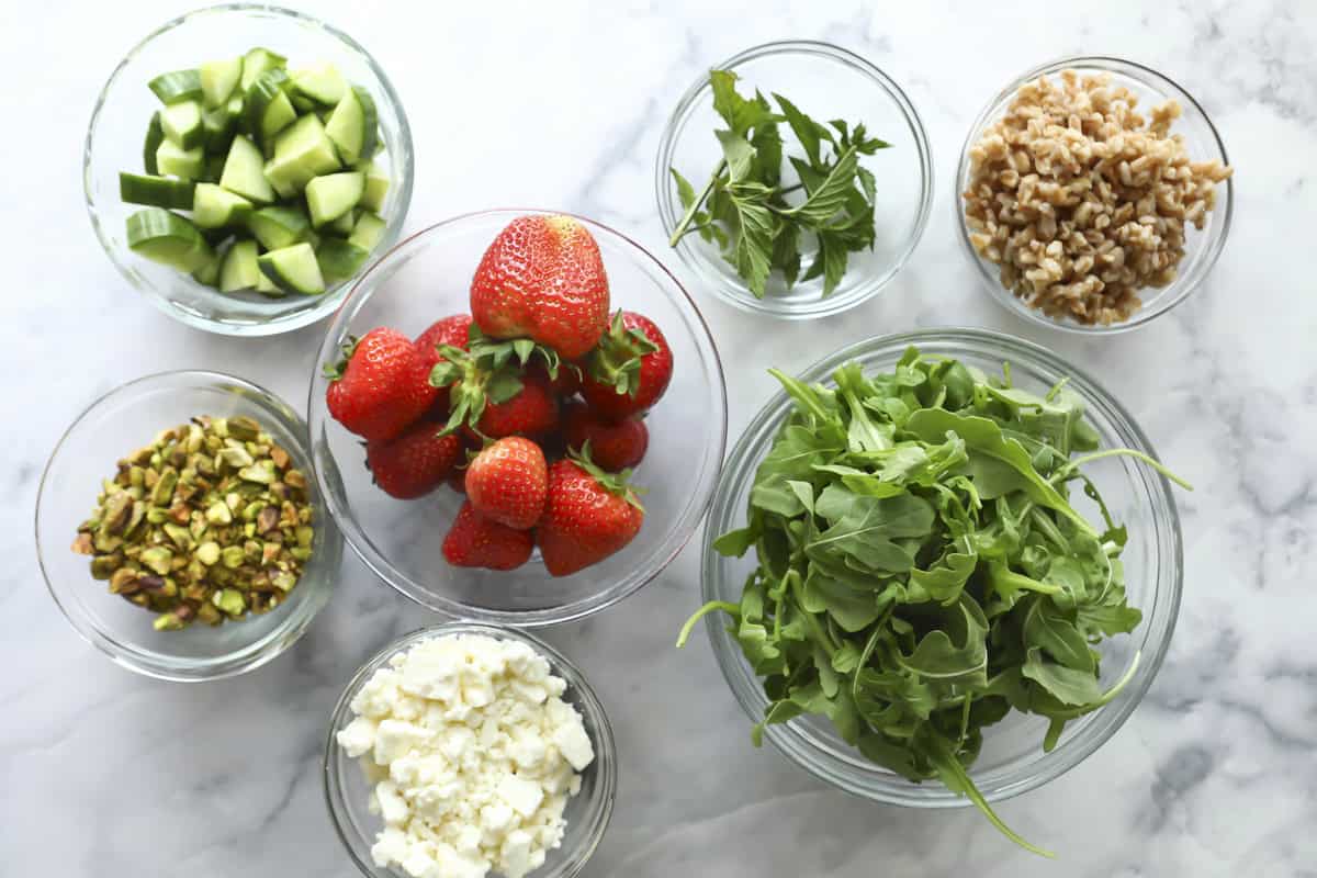 Bowls on a marble surface filled with fresh ingredients: diced cucumber, whole strawberries, walnuts, chopped herbs, arugula, crumbled feta cheese, and chopped pistachios.