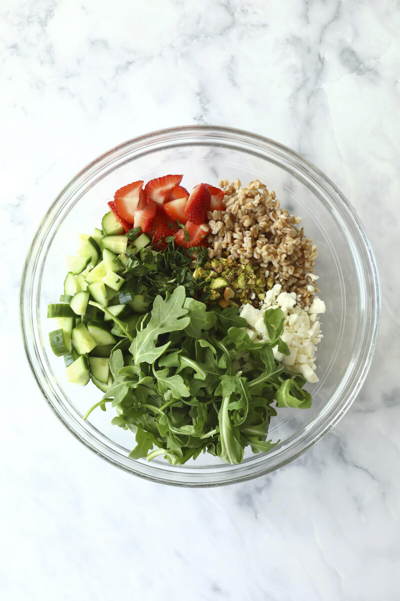 A glass bowl containing arugula, chopped cucumber, sliced strawberries, crumbled cheese, cooked grains, chopped herbs, and pistachios on a marble surface.