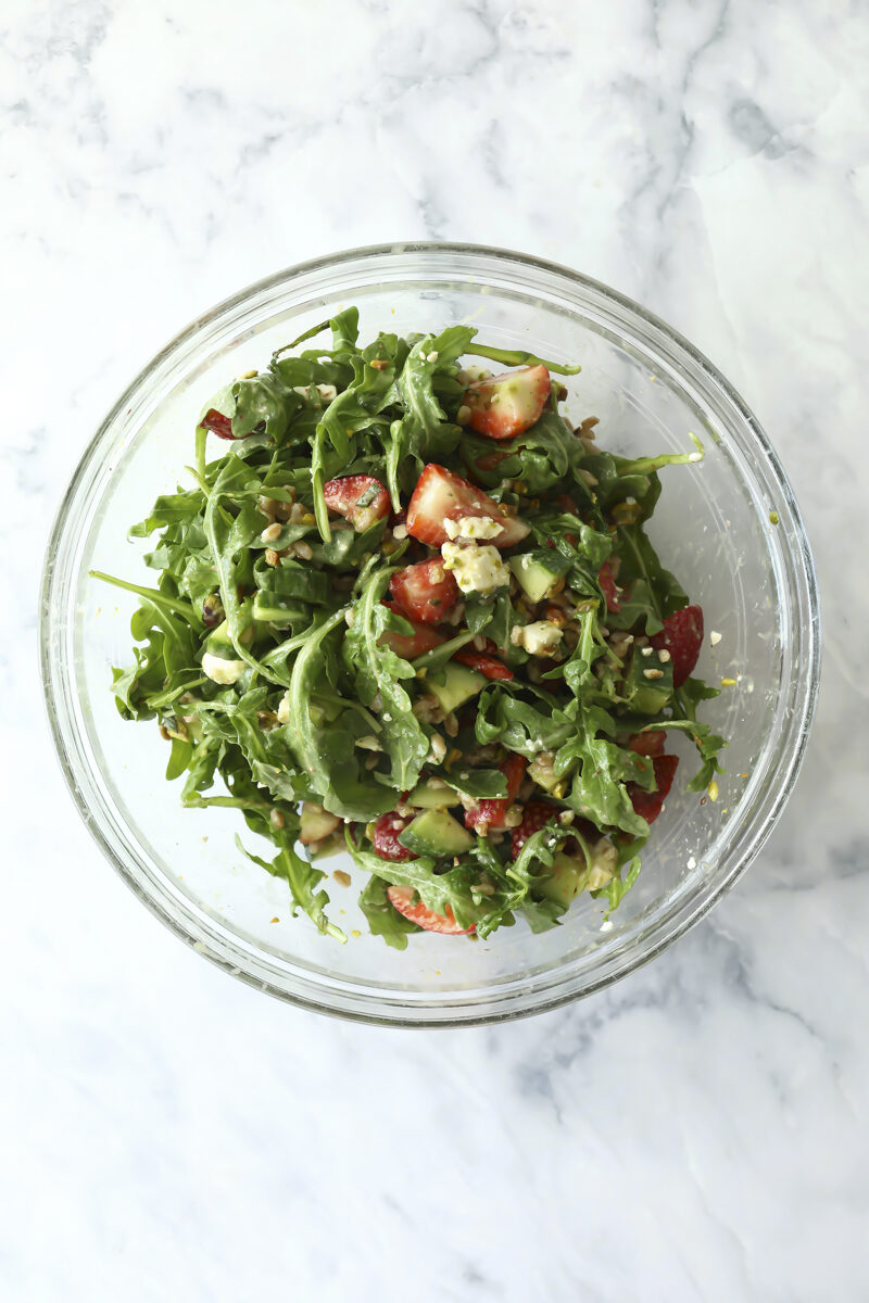 A glass bowl filled with a fresh salad containing arugula, sliced strawberries, cucumber, and seeds on a light marble surface.