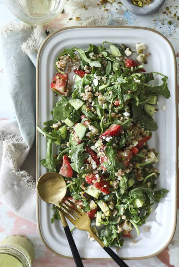 A rectangular white platter filled with a perfect summer salad of arugula, strawberries, cucumber, farro, feta cheese, and pistachios. Two serving utensils rest on the platter beside this perfect summer salad.