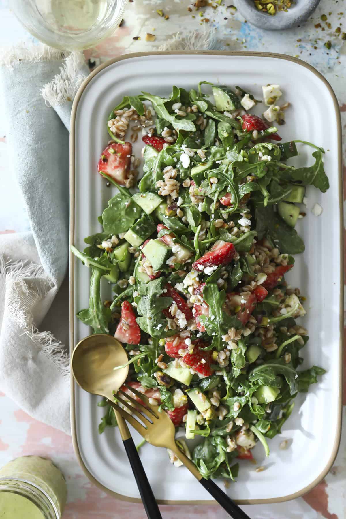 A rectangular white platter holds a fresh salad with arugula, strawberries, cucumber, grains, and feta cheese, garnished with seeds. Two salad servers rest on the side of the platter. A napkin and glass are nearby.