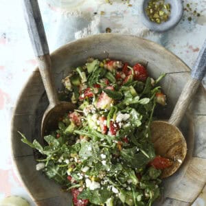 A wooden bowl filled with a fresh salad of greens, strawberries, cucumber, feta cheese, and nuts, with wooden salad servers. Glasses of white wine and small bowls of cheese and nuts are nearby on a light, textured surface.