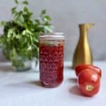 A jar of homemade tomato jam sits on a light surface, with two fresh tomatoes beside it. In the background, there is a bunch of leafy herbs in a glass and a gold-colored pitcher.