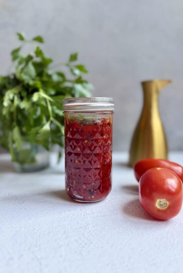A jar of homemade tomato jam sits on a light surface, with two fresh tomatoes beside it. In the background, there is a bunch of leafy herbs in a glass and a gold-colored pitcher.