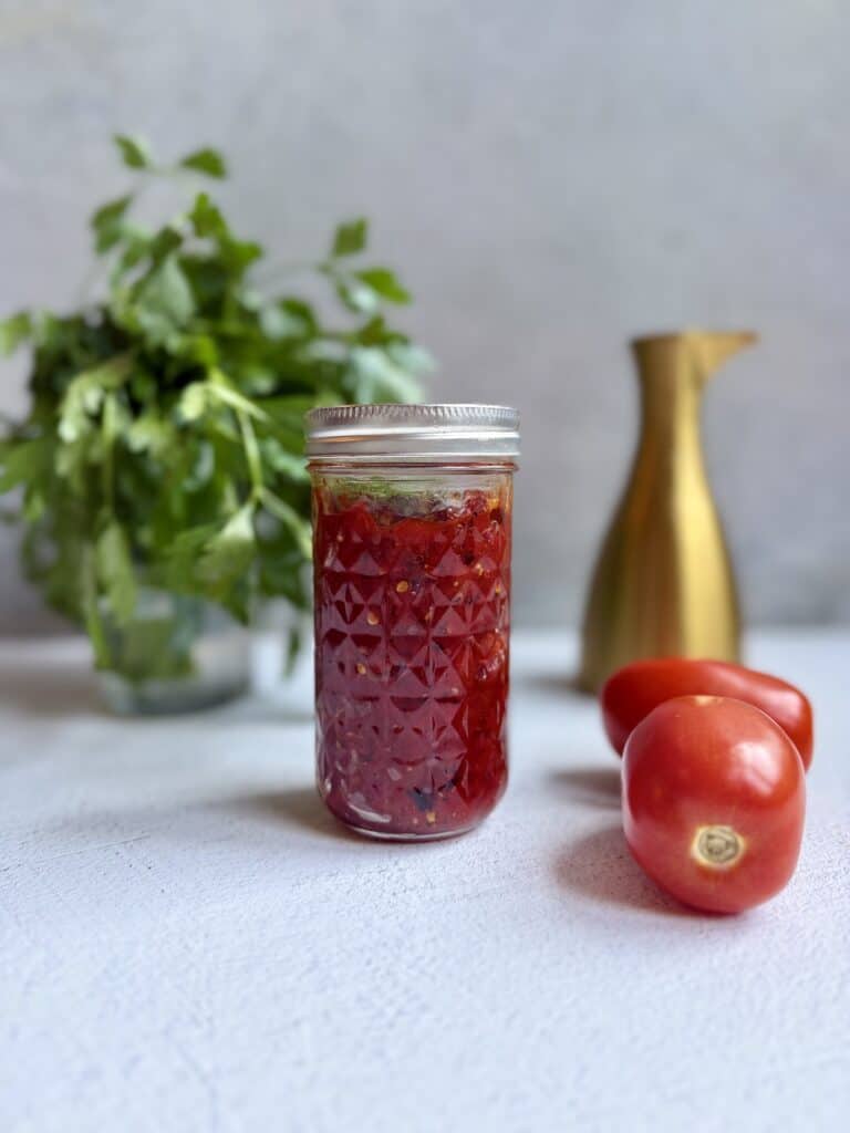 A jar of homemade tomato jam sits on a light surface, with two fresh tomatoes beside it. In the background, there is a bunch of leafy herbs in a glass and a gold-colored pitcher.