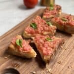Slices of toasted bread topped with fresh tomato spread and basil leaves are arranged on a wooden cutting board. A tomato and a gold bottle are blurred in the background.
