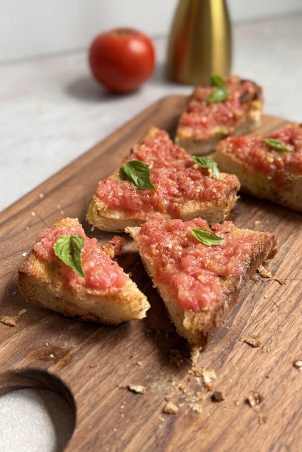 Slices of toasted bread topped with fresh tomato spread and basil leaves are arranged on a wooden cutting board. A tomato and a gold bottle are blurred in the background.