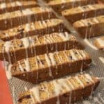 Rows of iced pumpkin biscotti are arranged on a baking sheet, each piece drizzled with a glaze. The biscotti are golden brown and studded with pieces of fruit or nuts, sitting on a silicone baking mat.