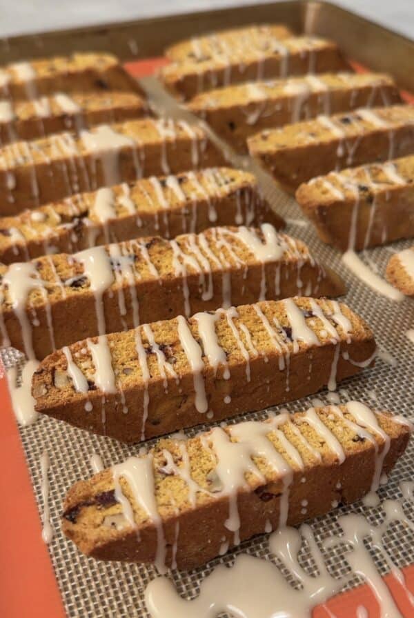 Rows of iced pumpkin biscotti are arranged on a baking sheet, each piece drizzled with a glaze. The biscotti are golden brown and studded with pieces of fruit or nuts, sitting on a silicone baking mat.