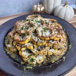 Baked delicata squash stack topped with melted cheese and chopped herbs, arranged in a stack. Decorative white and gold pumpkins are in the background on a wooden surface.