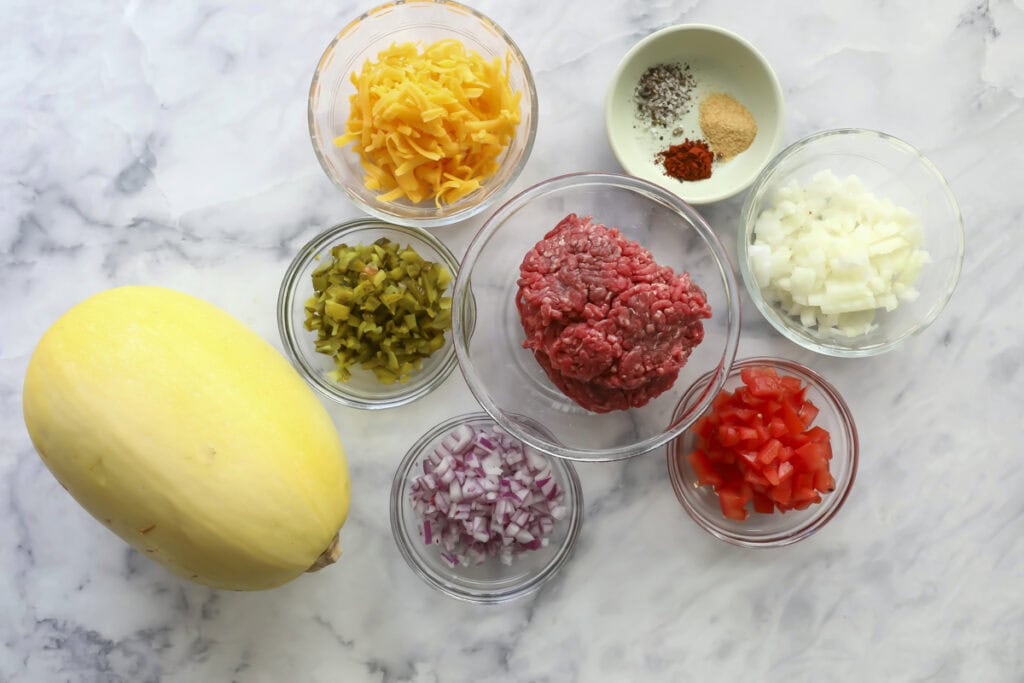 A whole spaghetti squash, ground beef, shredded cheese, chopped onion, red onion, diced tomato, chopped pickles, and seasoning are arranged in bowls on a marble countertop.
