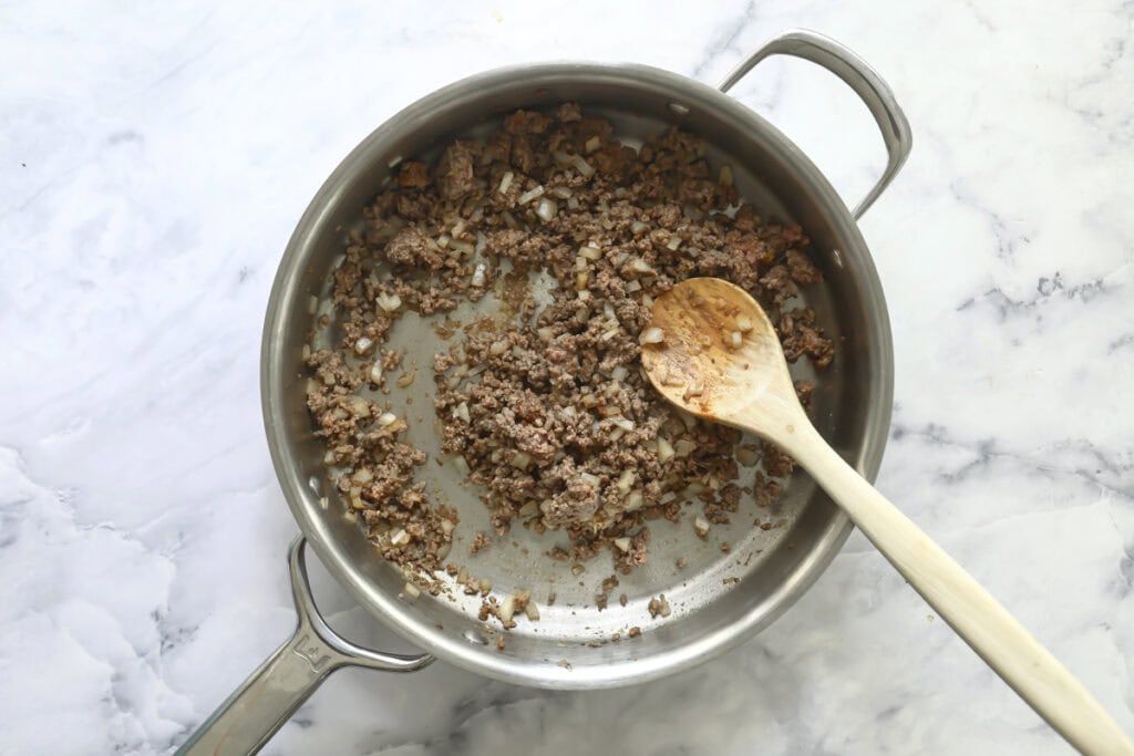 Ground meat and chopped onions being cooked in a stainless steel pan, stirred with a wooden spoon, on a white marble countertop.
