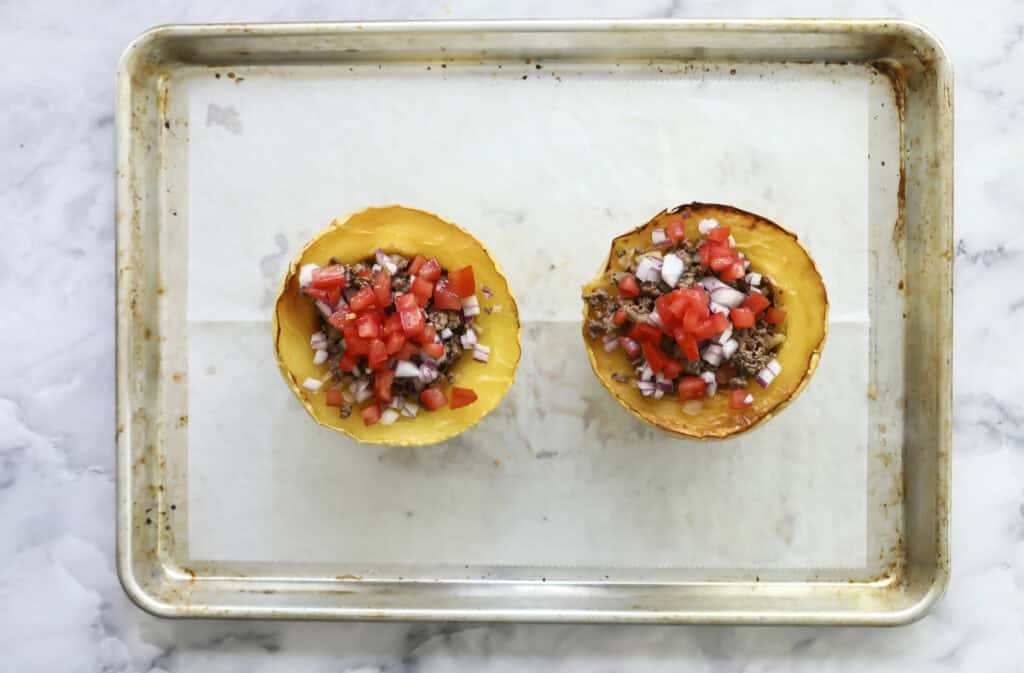 Two spaghetti squash halves on a parchment-lined baking tray, filled with a mixture of diced tomatoes, onions, and other vegetables. The tray sits on a white marble surface.
