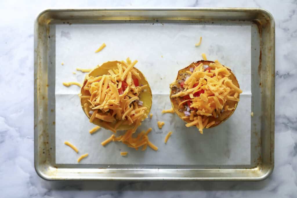 A baking sheet lined with parchment paper holds two halved stuffed peppers topped with shredded cheddar cheese, ready to be baked. The background is a white marble surface.