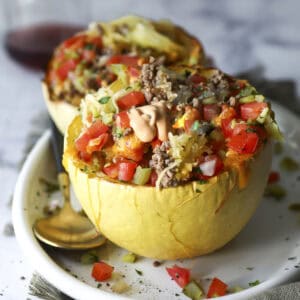 A halved Big Mac spaghetti squash stuffed with ground meat, diced tomatoes, onions, pickles, and drizzled with sauce sits on a white plate with a gold spoon, set on a cloth napkin.