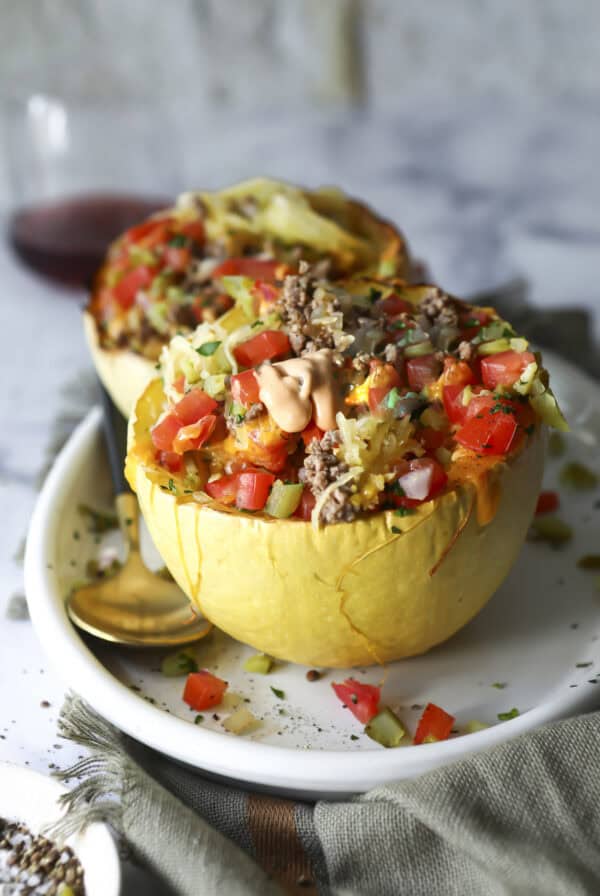 A halved Big Mac spaghetti squash stuffed with ground meat, diced tomatoes, onions, pickles, and drizzled with sauce sits on a white plate with a gold spoon, set on a cloth napkin.