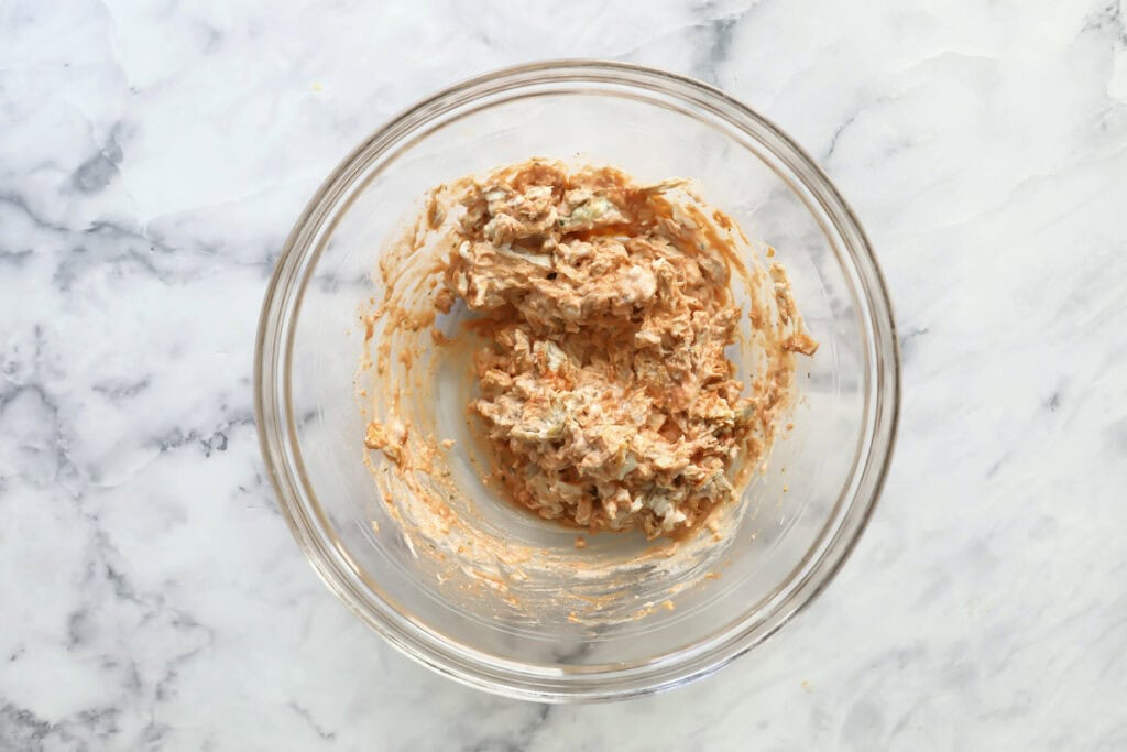 A clear glass bowl on a marble countertop contains a partially-mixed spread with a creamy, orange-tinted texture and visible chunks, likely a mixture of cream cheese and seasonings.