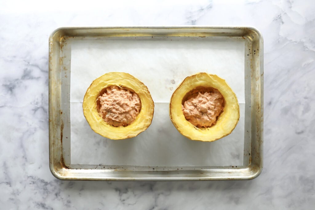 A baking sheet with two spaghetti squash halves filled with a creamy mixture, placed on parchment paper, set on a marble countertop.