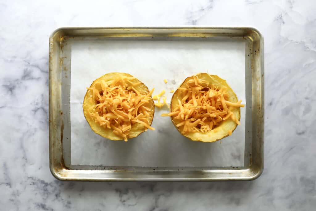 Two halved spaghetti squash filled with shredded cheese sit on a parchment-lined baking tray, ready for baking. The tray is on a white marble surface.