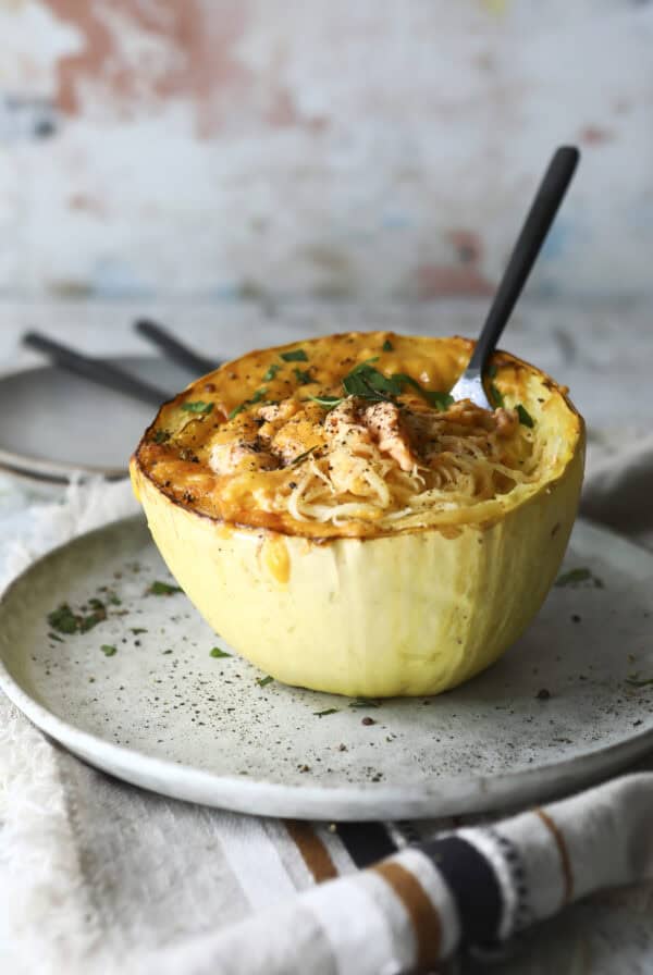 A baked spaghetti squash half filled with cheesy spaghetti and garnished with herbs sits on a ceramic plate, with a fork inserted, ready to eat.